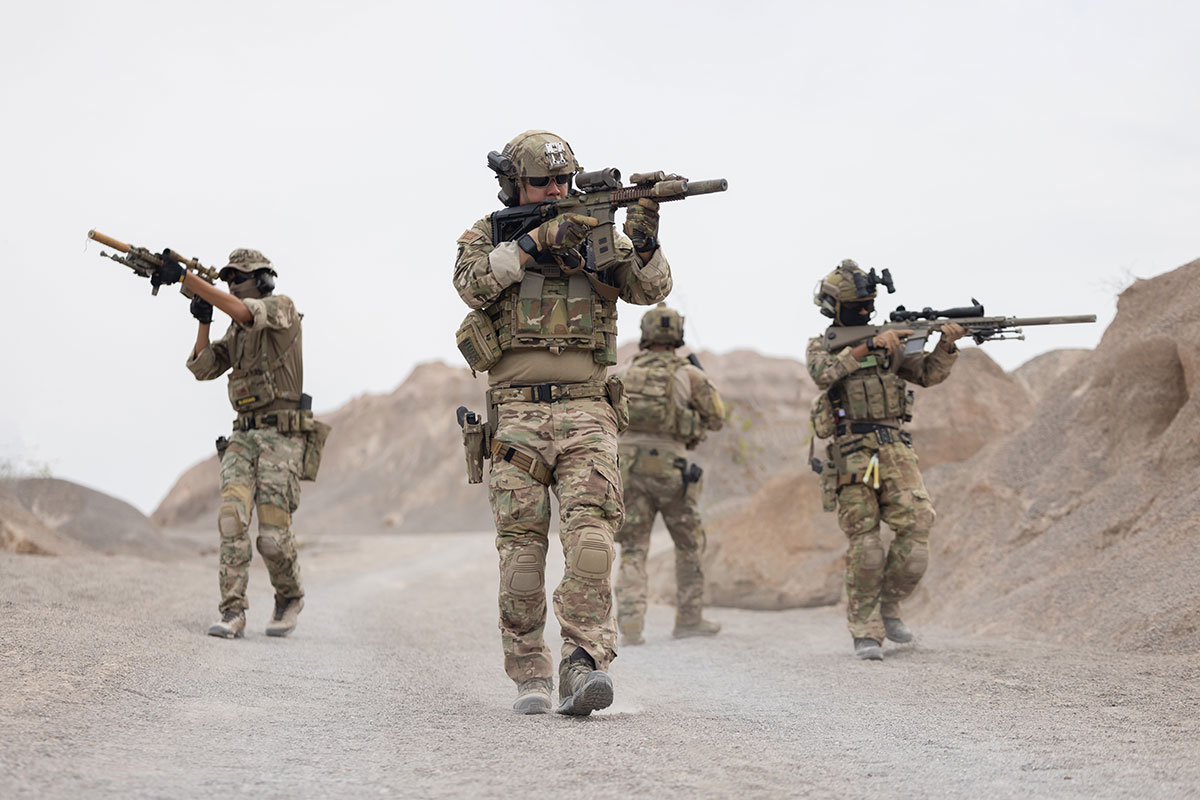 A group of soldiers in camouflage uniforms conducting a patrol or training exercise in a desert-like, rugged mountain environment.
